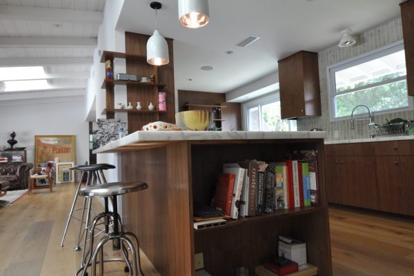 Open-concept kitchen featuring a walnut island with built-in shelving, industrial-style metal bar stools, white pendant lights, wide-plank wood flooring, and matching walnut cabinetry with large windows.