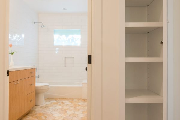 Hallway view into a modern bathroom with glossy honey-onyx hexagon flooring, white subway tile walls, wood vanity, and adjacent built-in linen cabinet with open shelving.