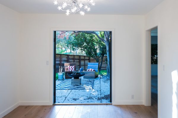 Bright interior room with wide sliding doors opening to a cozy outdoor seating area featuring modern chairs, patterned rug, fire bowl, and a fenced backyard with mature trees.