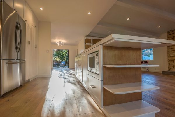 Modern kitchen with extra-long island, built-in microwave, open corner shelving, white cabinetry, stainless steel appliances, and wide-plank wood flooring extending toward a bright backyard entry.