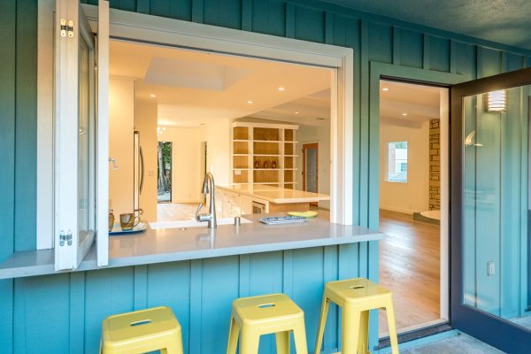 Indoor–outdoor kitchen passthrough with teal board-and-batten exterior, quartz countertop bar, open bi-fold window, and yellow metal bar stools at a remodeled midcentury home.
