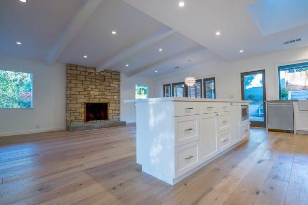 Open-concept living room and kitchen remodel featuring wide-plank oak flooring, a large white island with shaker cabinetry, vaulted ceilings with beams, and a stone fireplace—Cozy Canopy House by Driftwood Studio Inc.