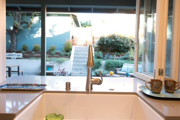 Kitchen pass-through window with farmhouse sink overlooking a terraced backyard with outdoor seating, lush landscaping, and concrete steps — Cozy Canopy House remodel by Driftwood Studio Inc.