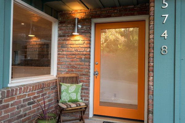 Warm and inviting front entry with orange modern door, brick exterior, teal board-and-batten siding, metal house numbers, and small seating area — Cozy Canopy House by Driftwood Studio Inc.