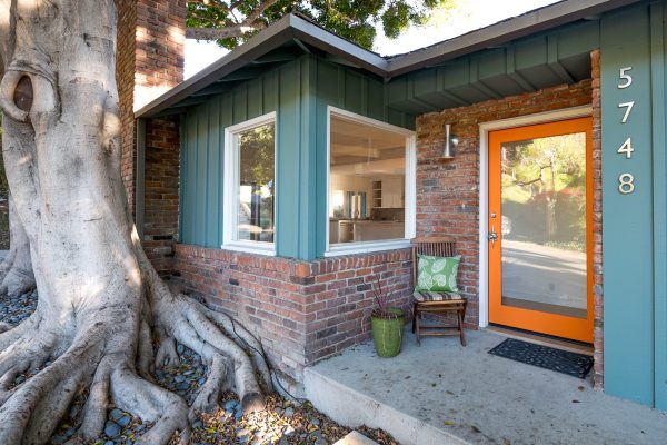 Front entry of teal board-and-batten home with orange door, brick exterior, and large sculptural tree roots beside the porch.