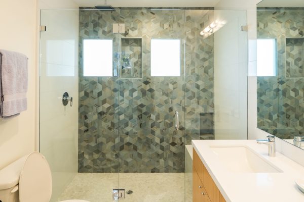 Modern bathroom with geometric tile shower, frameless glass enclosure, white quartz vanity, natural wood cabinetry, and hexagon mosaic floor — Cozy Canopy House remodel by Driftwood Studio Inc.