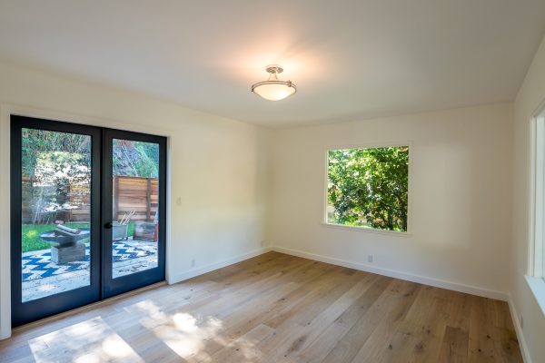 Bright bedroom with wide-plank oak floors, large picture window, black-framed French doors, and views of a private patio and lush backyard — Cozy Canopy House by Driftwood Studio Inc.